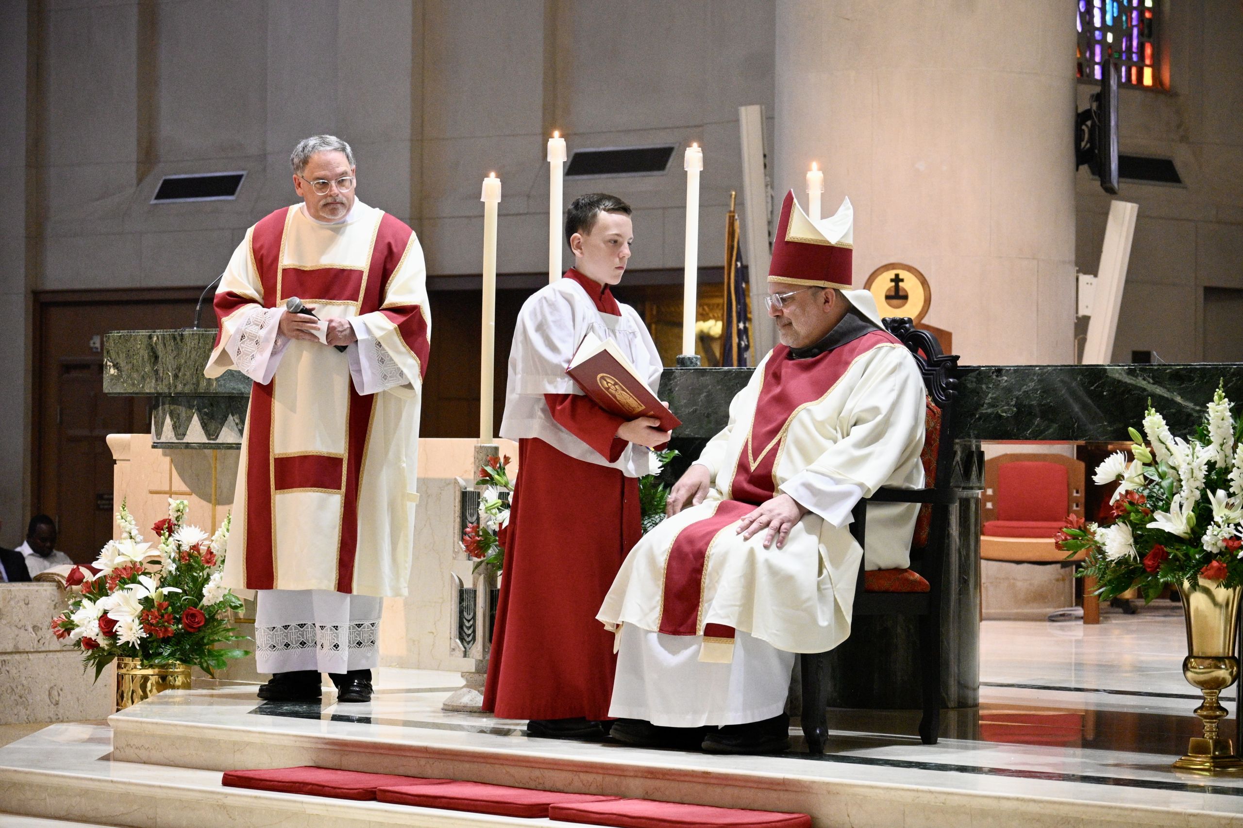 A bishop in red and white liturgical vestments sits at a marble altar as an acolyte reads from a red book nearby, with tall candles and floral arrangements around them in a church.