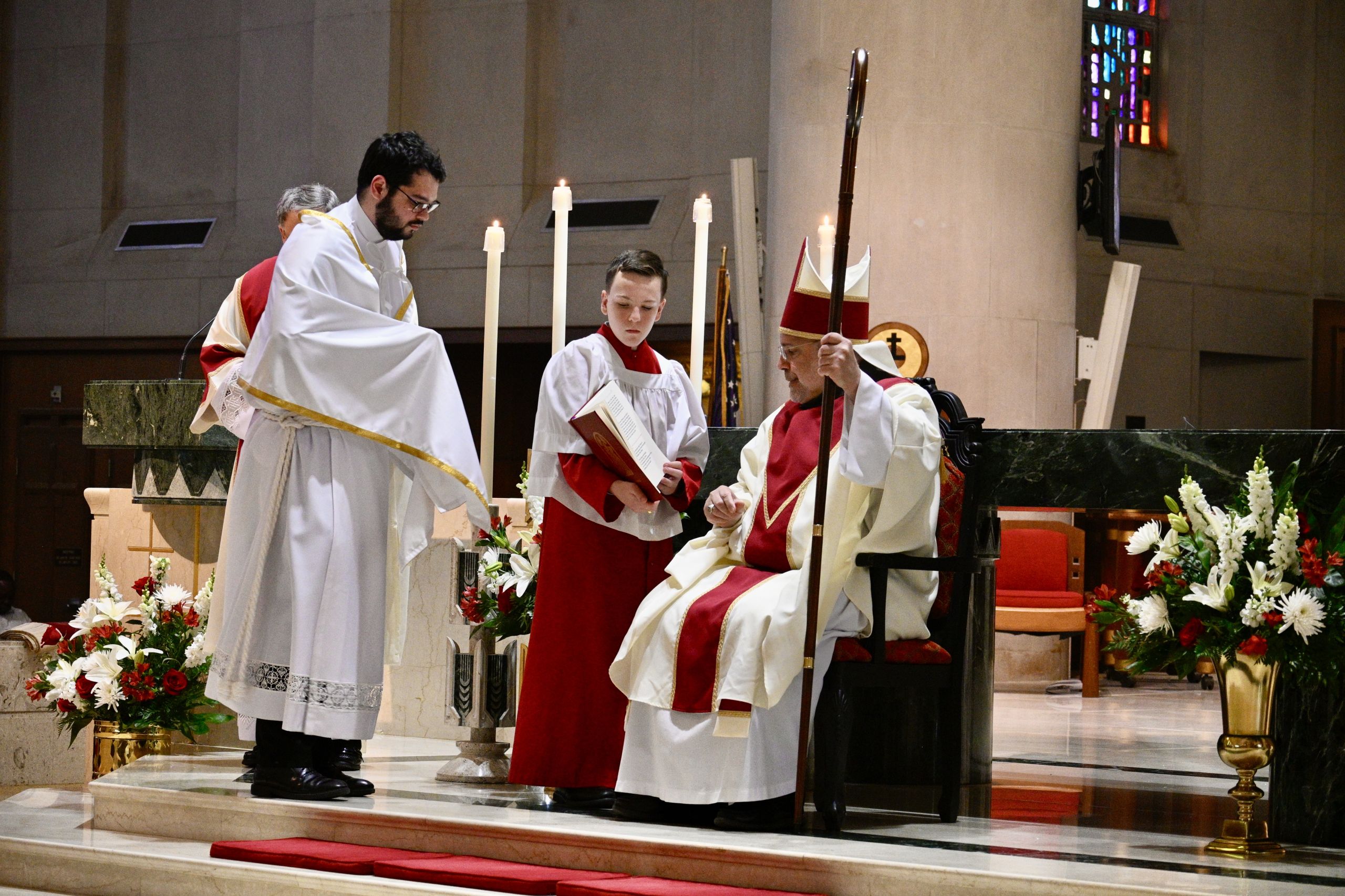 Clergy in white and red vestments perform a liturgical rite at the altar with tall candles nearby.