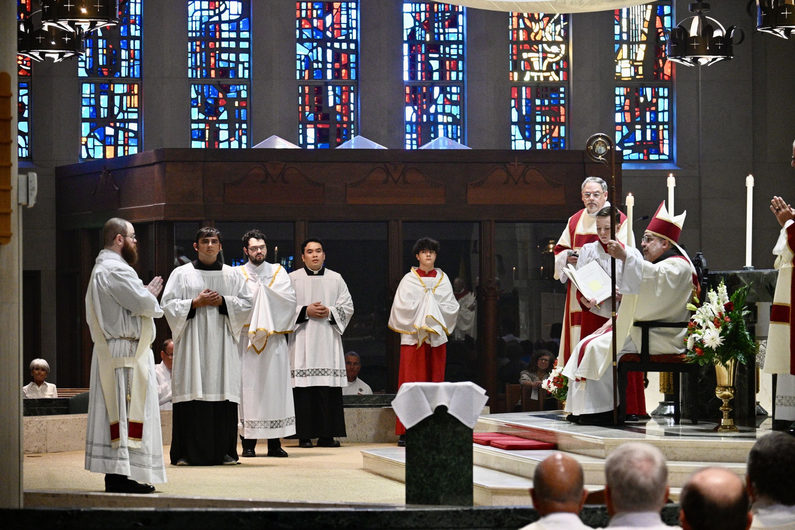 Clergy in white robes stand at a church altar during a ceremony, with a bishop in red and gold leading the service and stained-glass windows behind them.