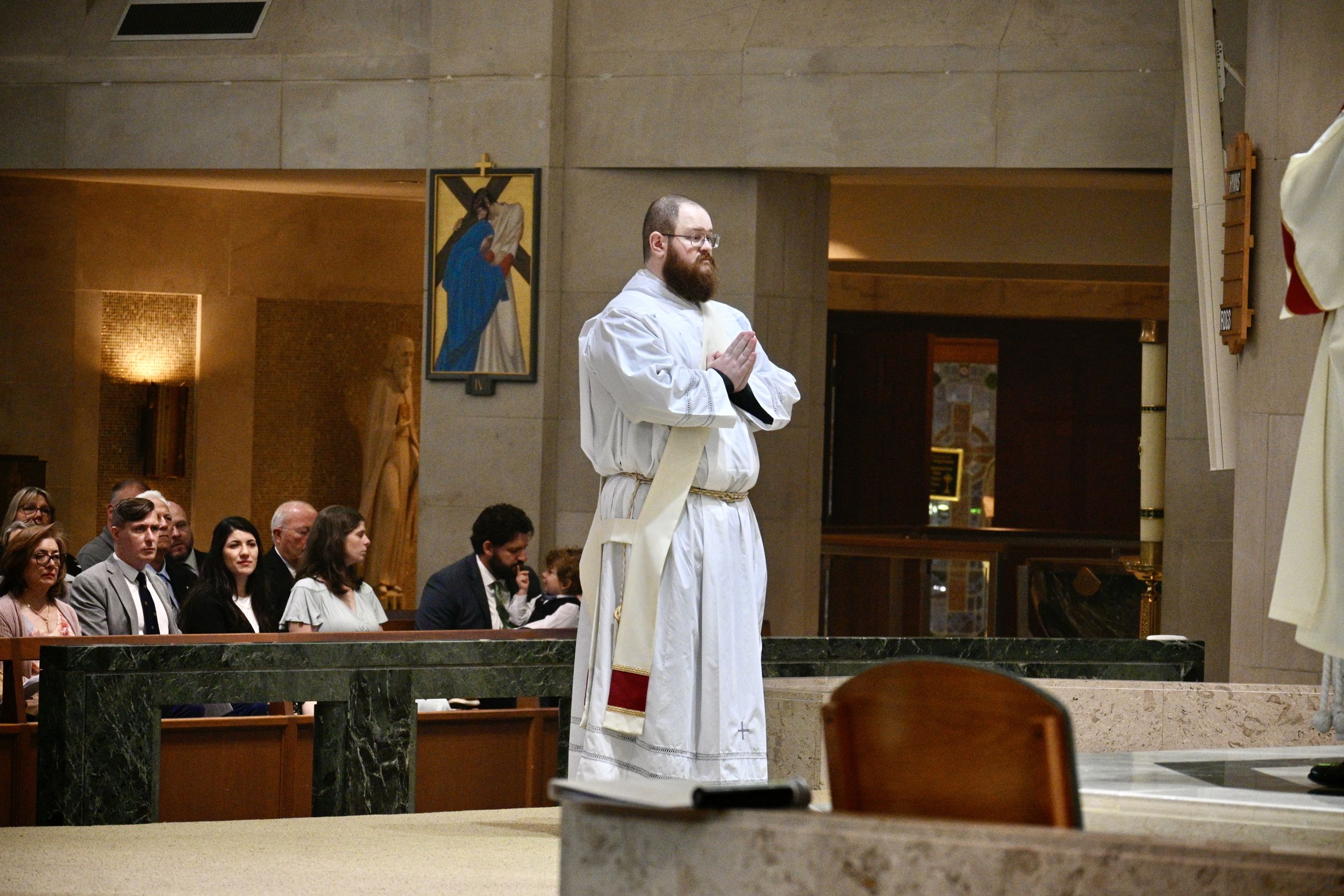 Bearded priest in white robes with hands joined in prayer at the front of a church, congregation seated to the left.