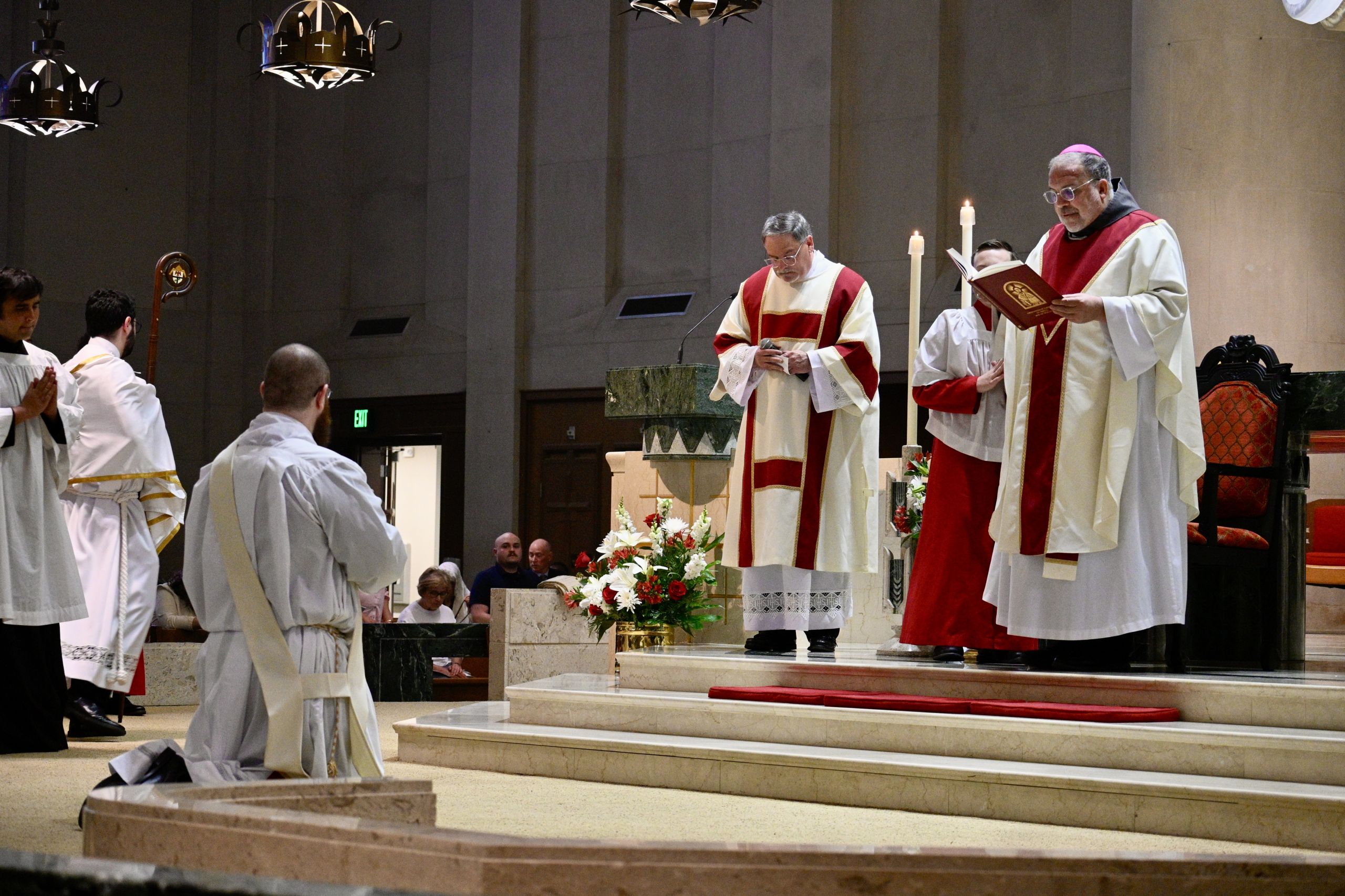 Priests in red-and-white vestments reading prayers at a marble altar during Mass, with flowers nearby.