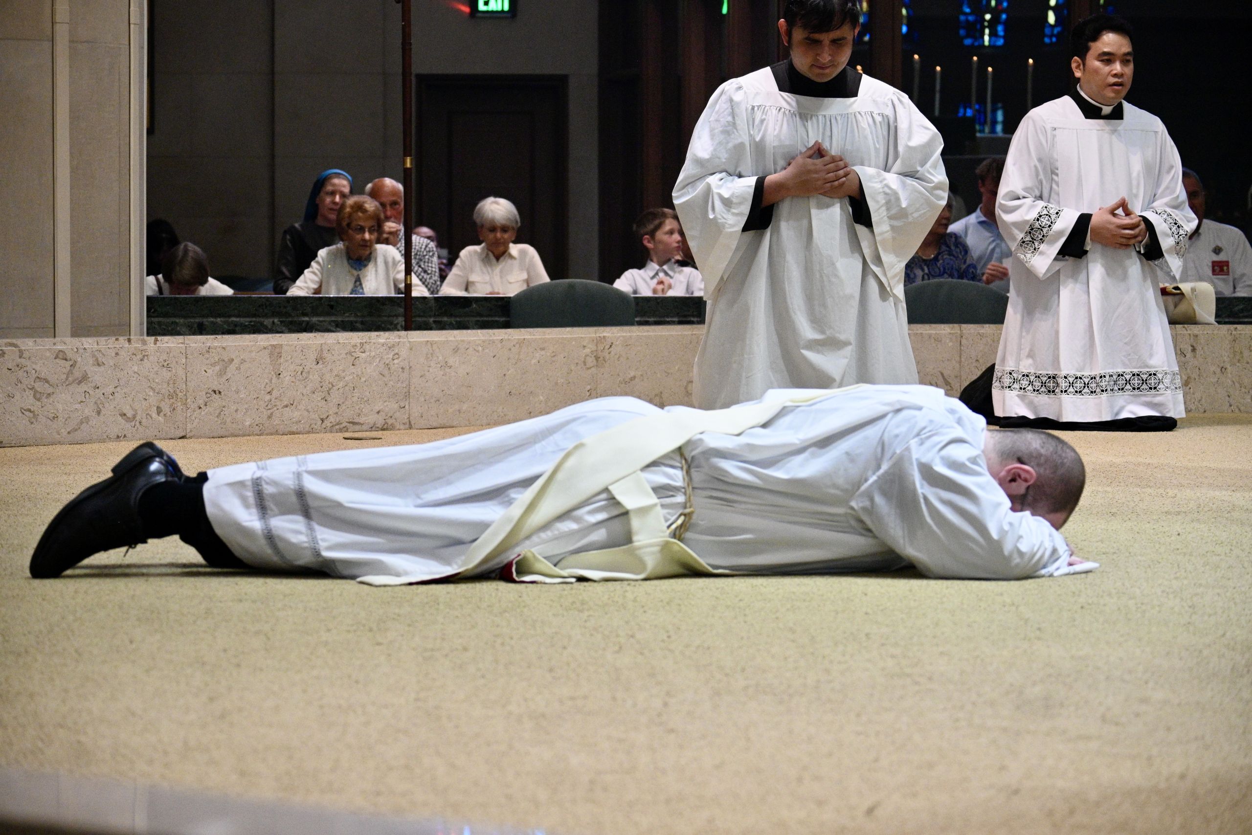 Funeral scene in a church: two priests in white robes stand with hands folded near a body wrapped in a white shroud on the floor behind a marble railing.