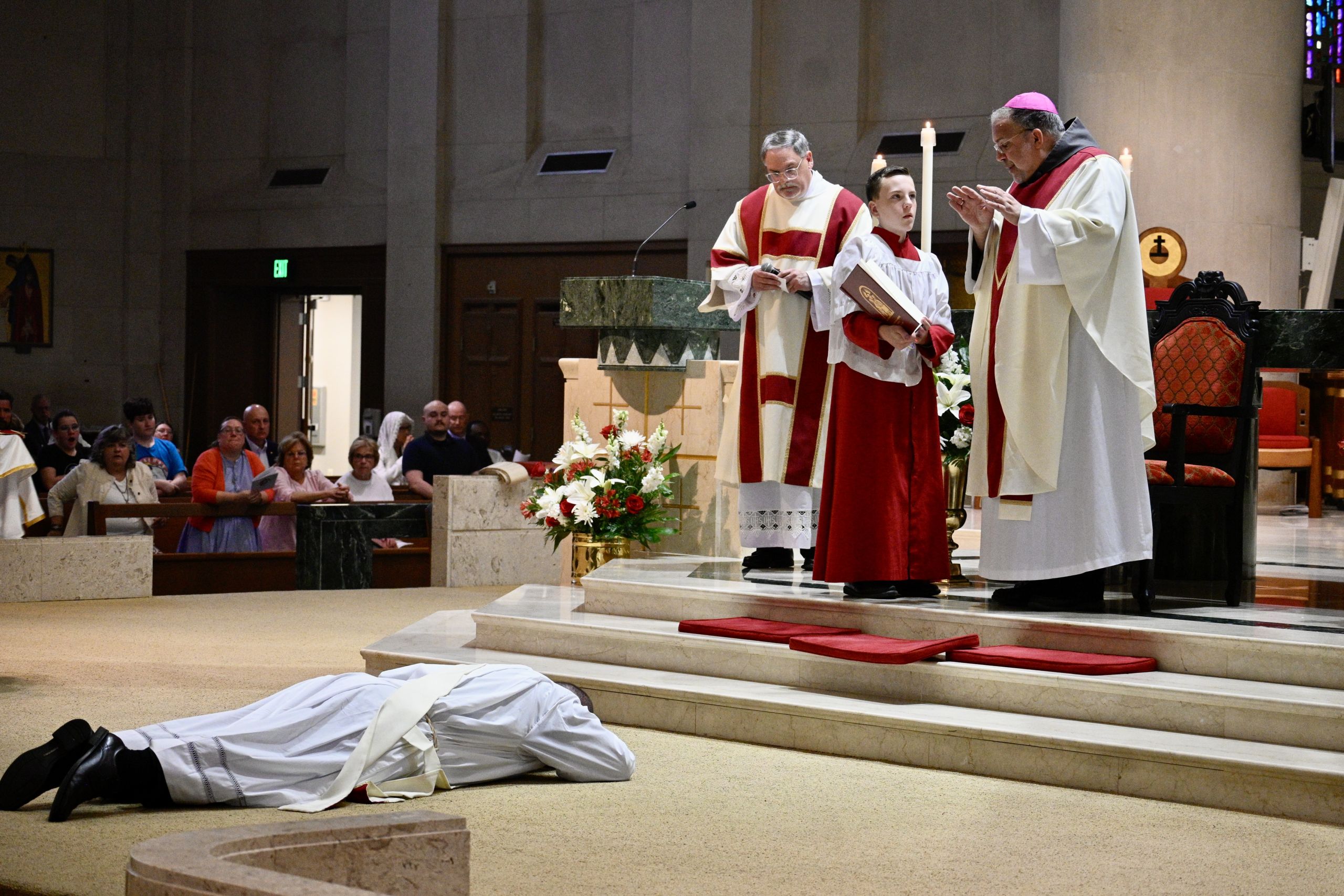 Clergy in red and white robes perform a service as a lifeless body wrapped in white shroud lies on the church steps in front of the altar area.