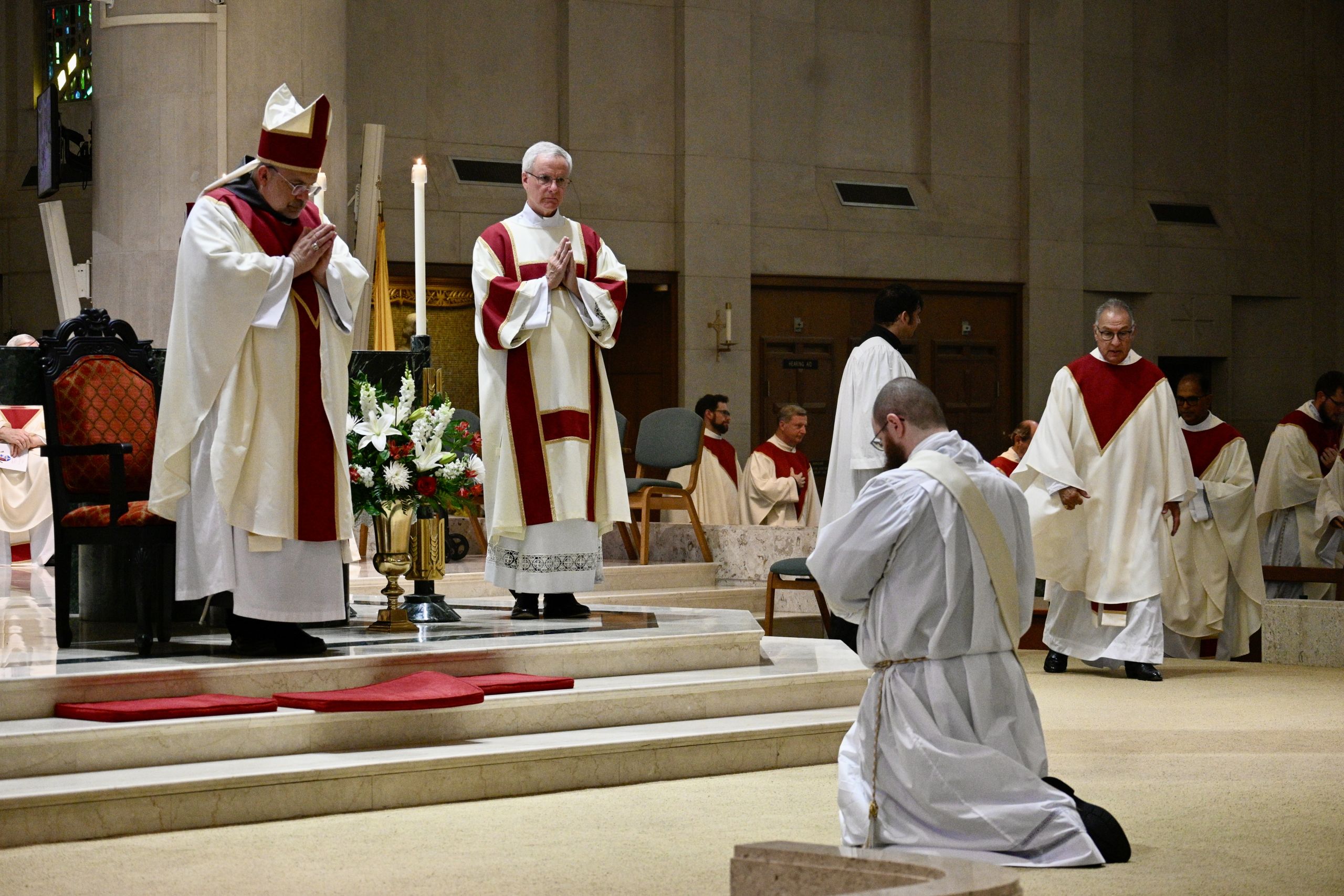 Catholic clergy in white and red vestments pray at the altar during a liturgical ceremony, with flowers nearby and kneeling assistants in the background.