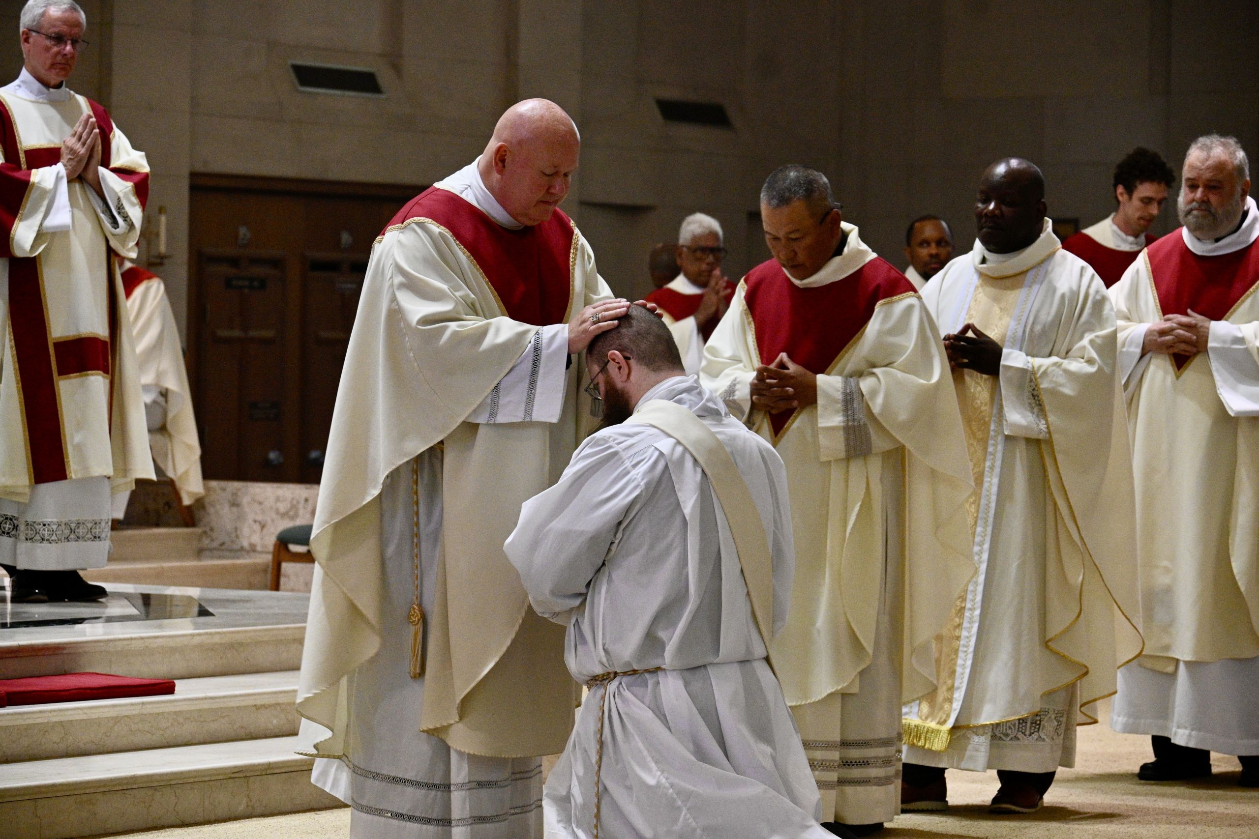 Priest places hands on kneeling man during a church ceremony, surrounded by clergy in white robes with red stoles.