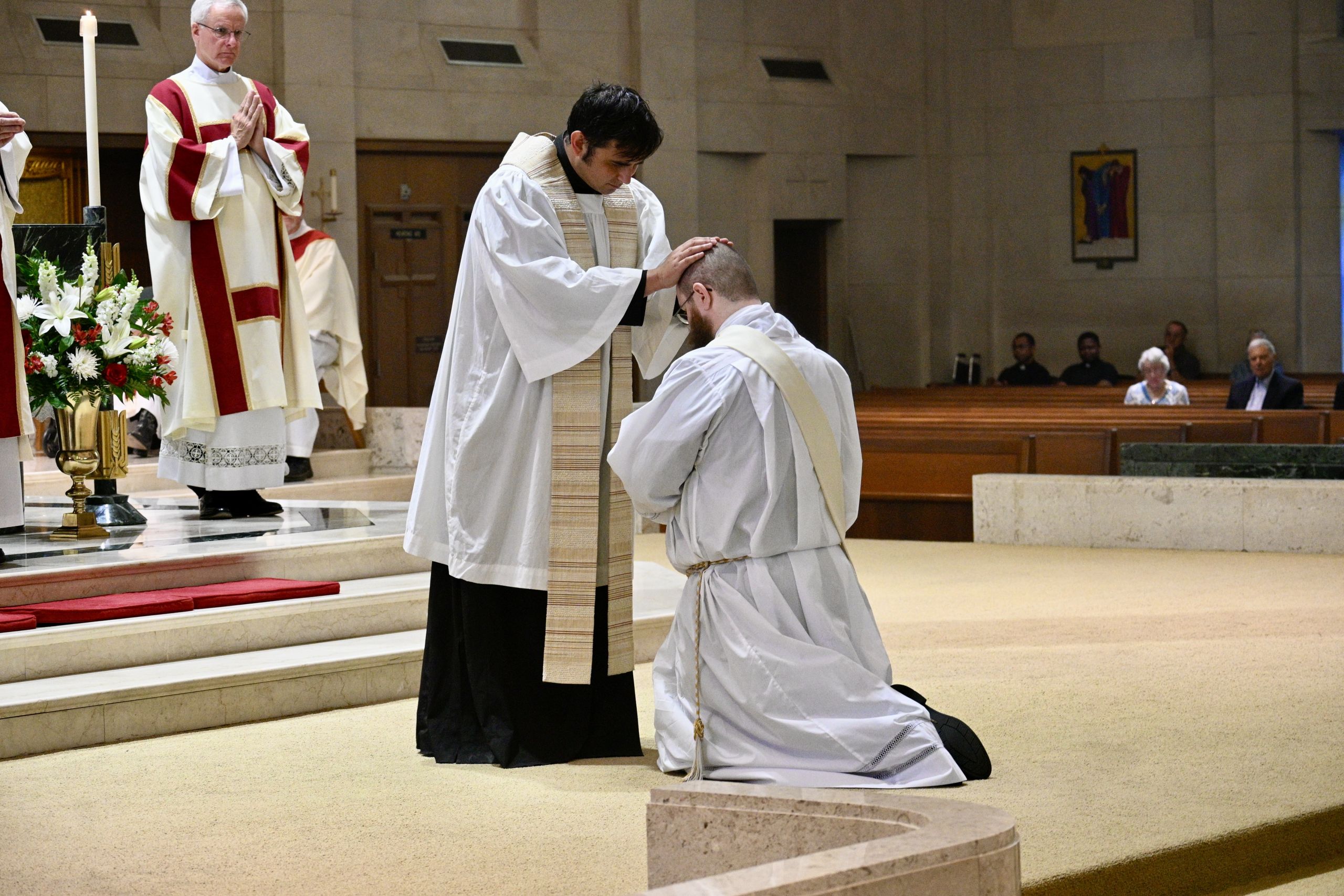 Priest places hand on kneeling clergyman's head during a liturgical blessing at the altar in a church.