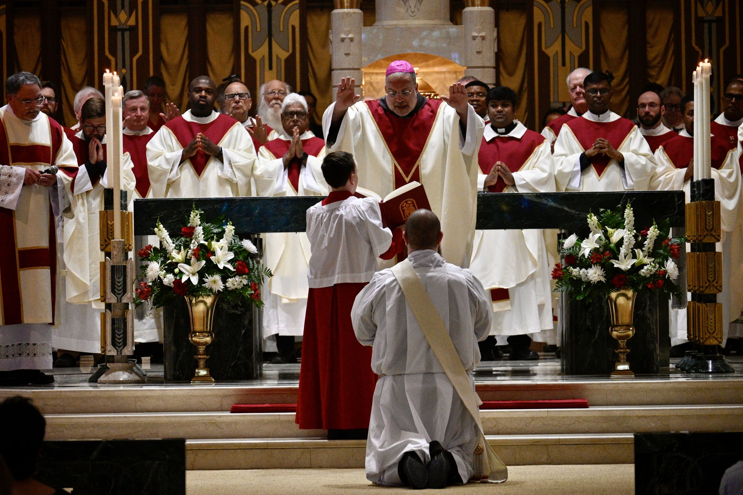 Catholic Mass: a bishop blesses a kneeling boy as priests in red and white vestments stand behind an ornate altar with flowers.