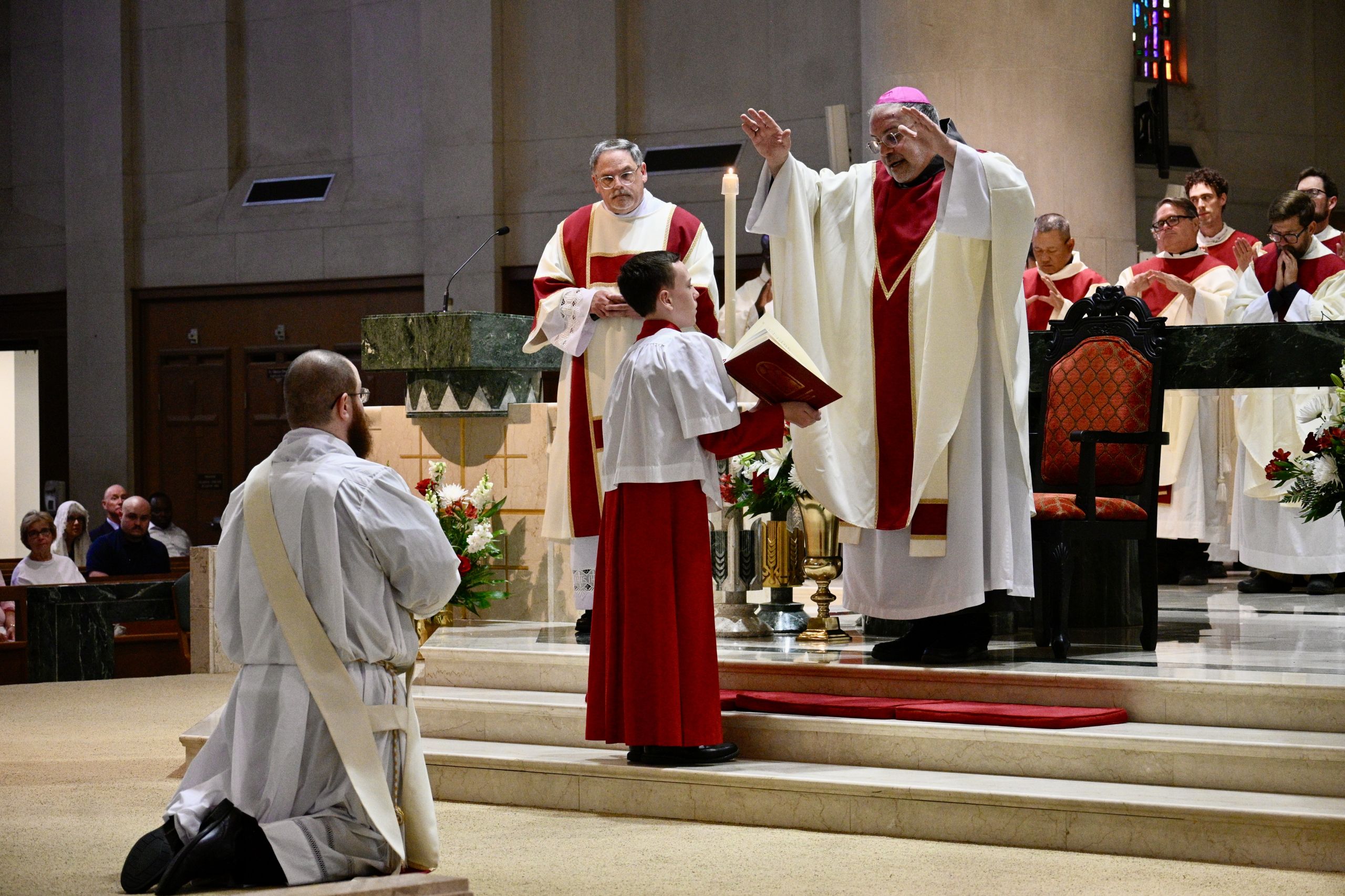 Catholic bishop raises hands in blessing over a kneeling boy in white and red vestments at the altar during a ceremony.