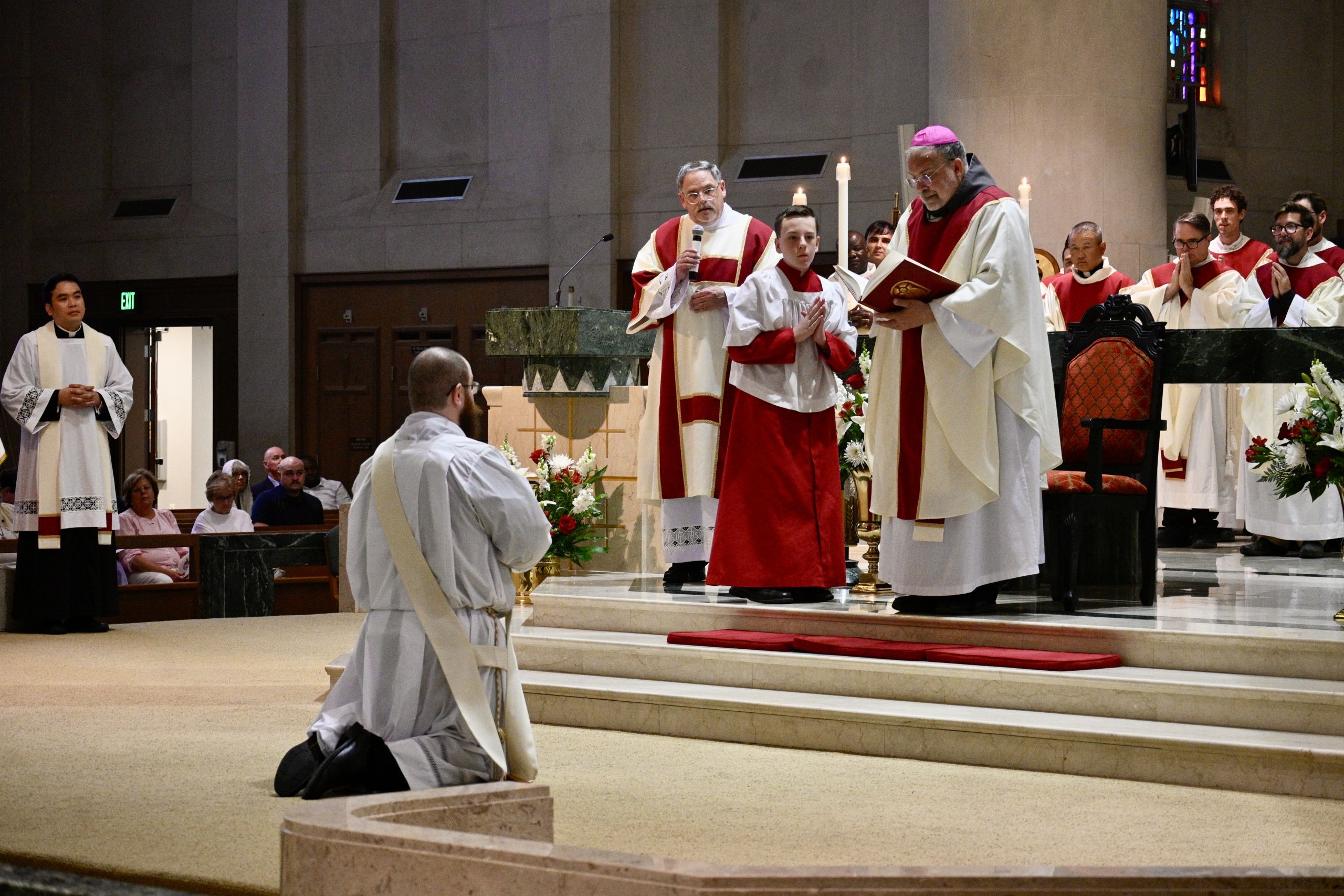 Clergy in red and white vestments lead a Mass at a marble altar, with a kneeling participant in the foreground.