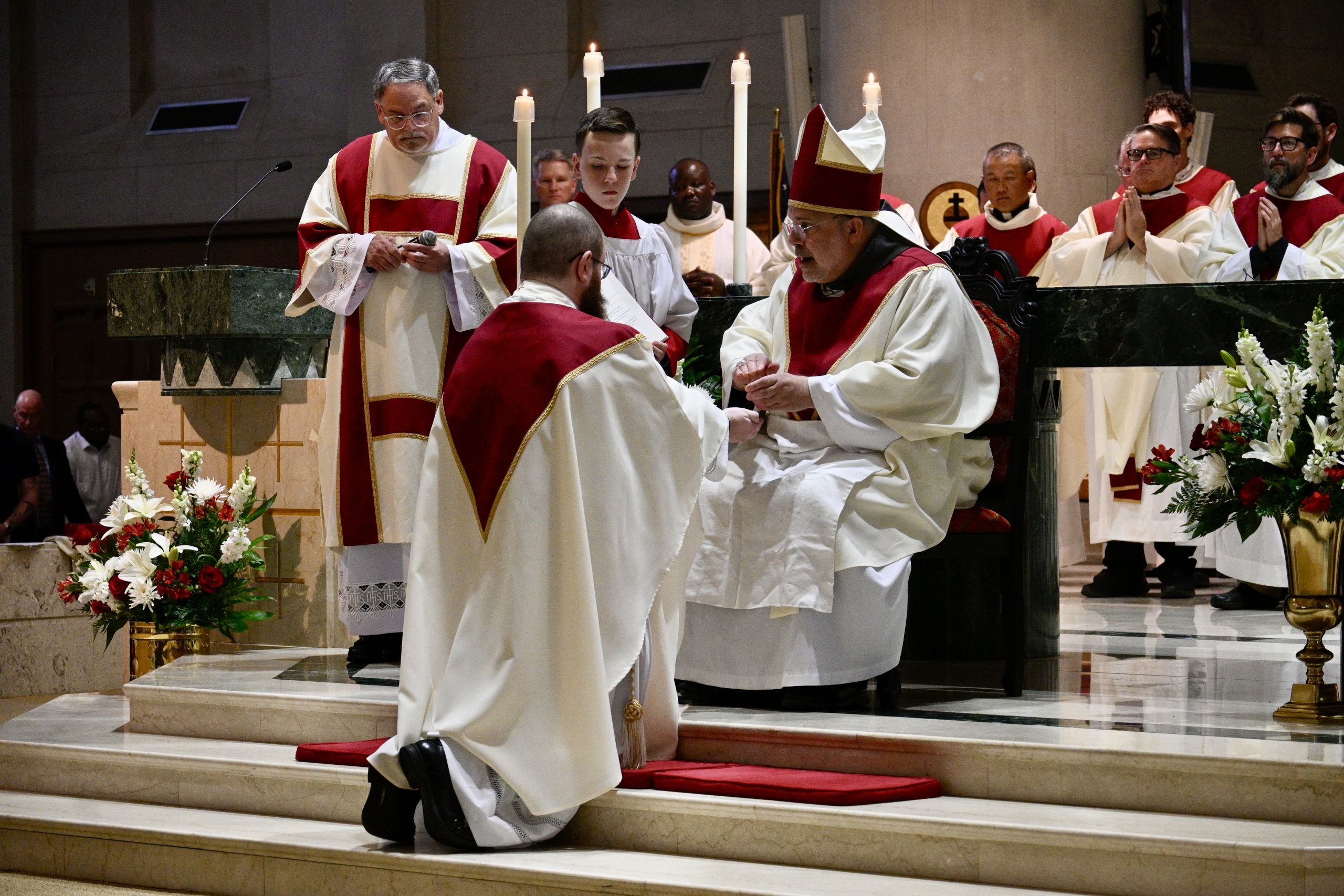 Priests in red and white liturgical robes conduct a ceremony at a marble altar with tall candles and floral arrangements surrounding them.