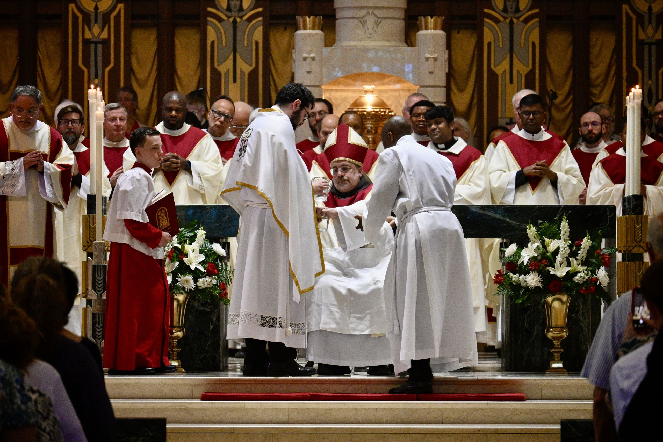 Priests in red and white vestments surround an ornate altar as a bishop in a red mitre distributes communion.