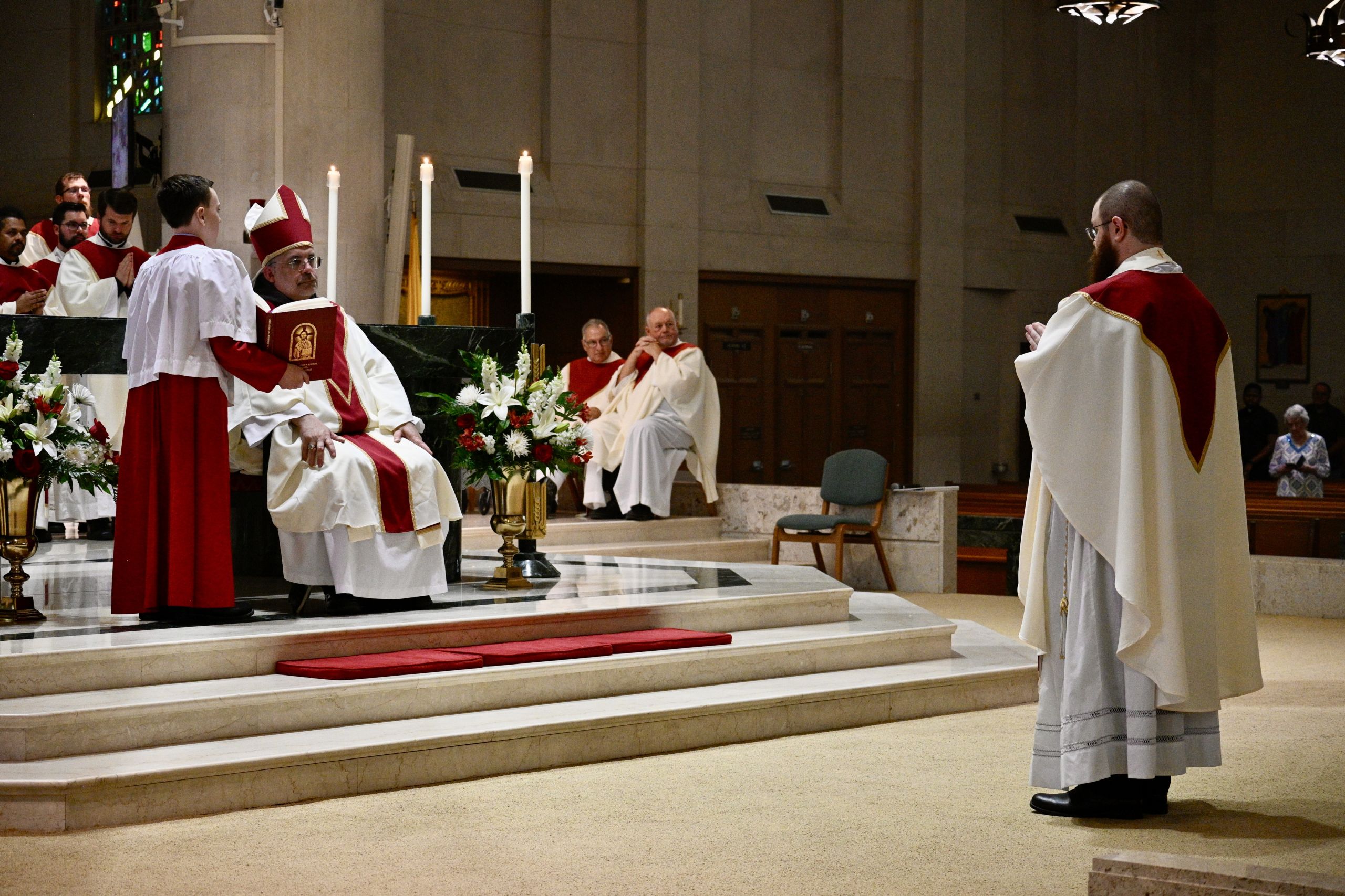 Bishop in red and white vestments seated at the altar, holding a red liturgical book beside candles and flowers.