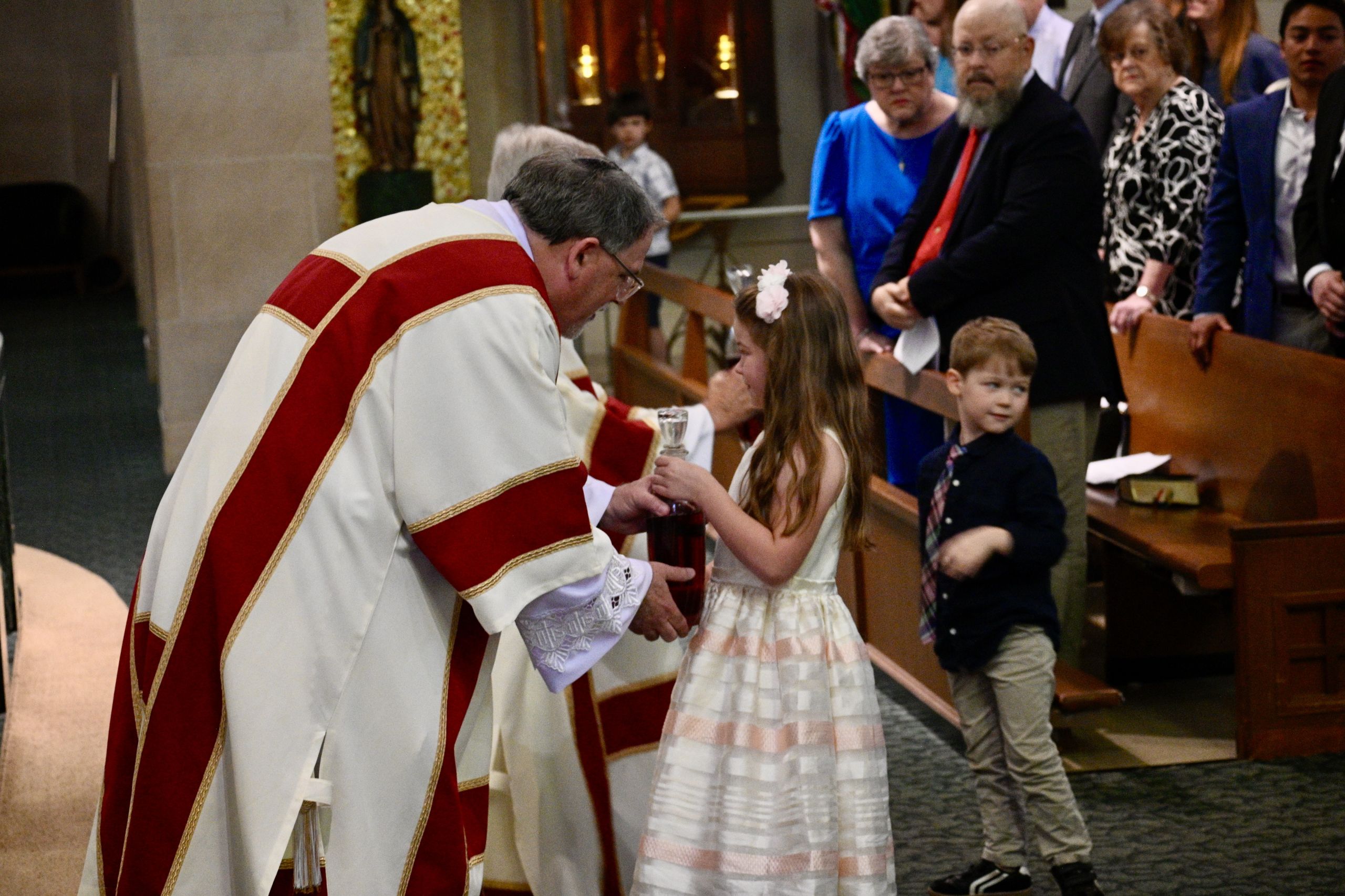 Priest in red-and-white vestments blesses a young girl at a church ceremony, holding a chalice.