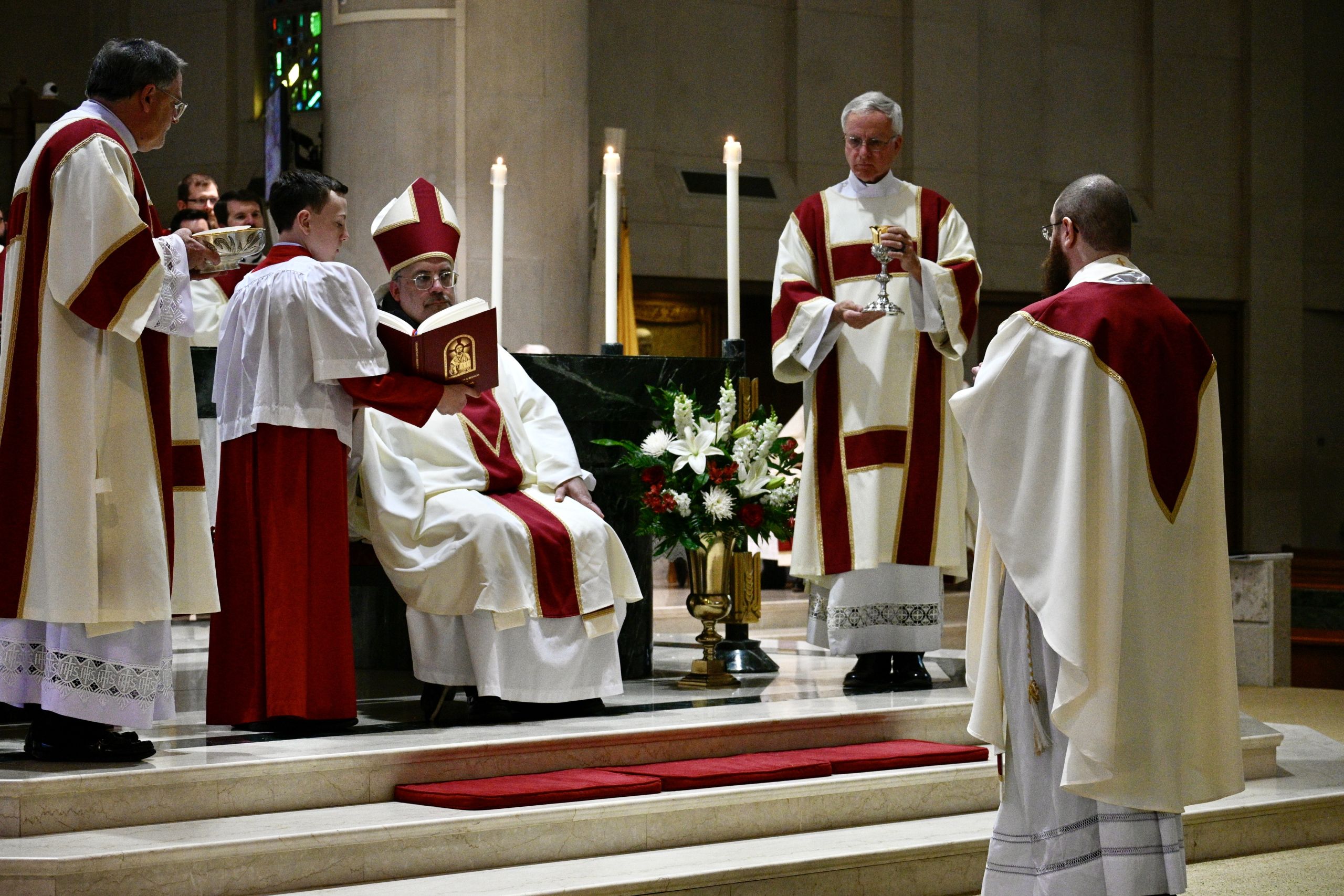 Clergy in red and white vestments perform a Catholic service at an altar with candles and flowers.
