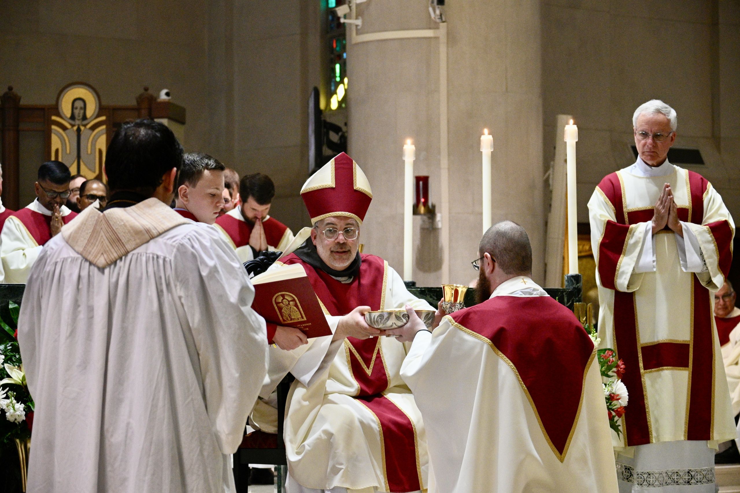 Catholic bishop in red and white vestments officiating at an altar with clergy and lit candles nearby.
