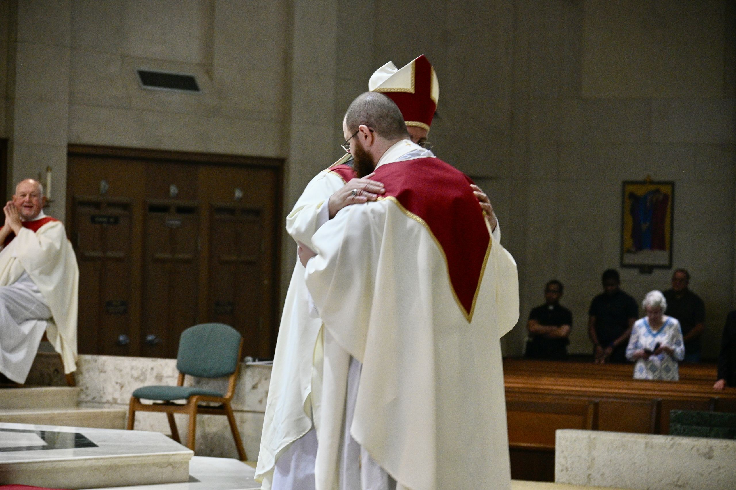 Two clergy members in white and red liturgical vestments hug on a church stage, with seated and standing spectators in the background.