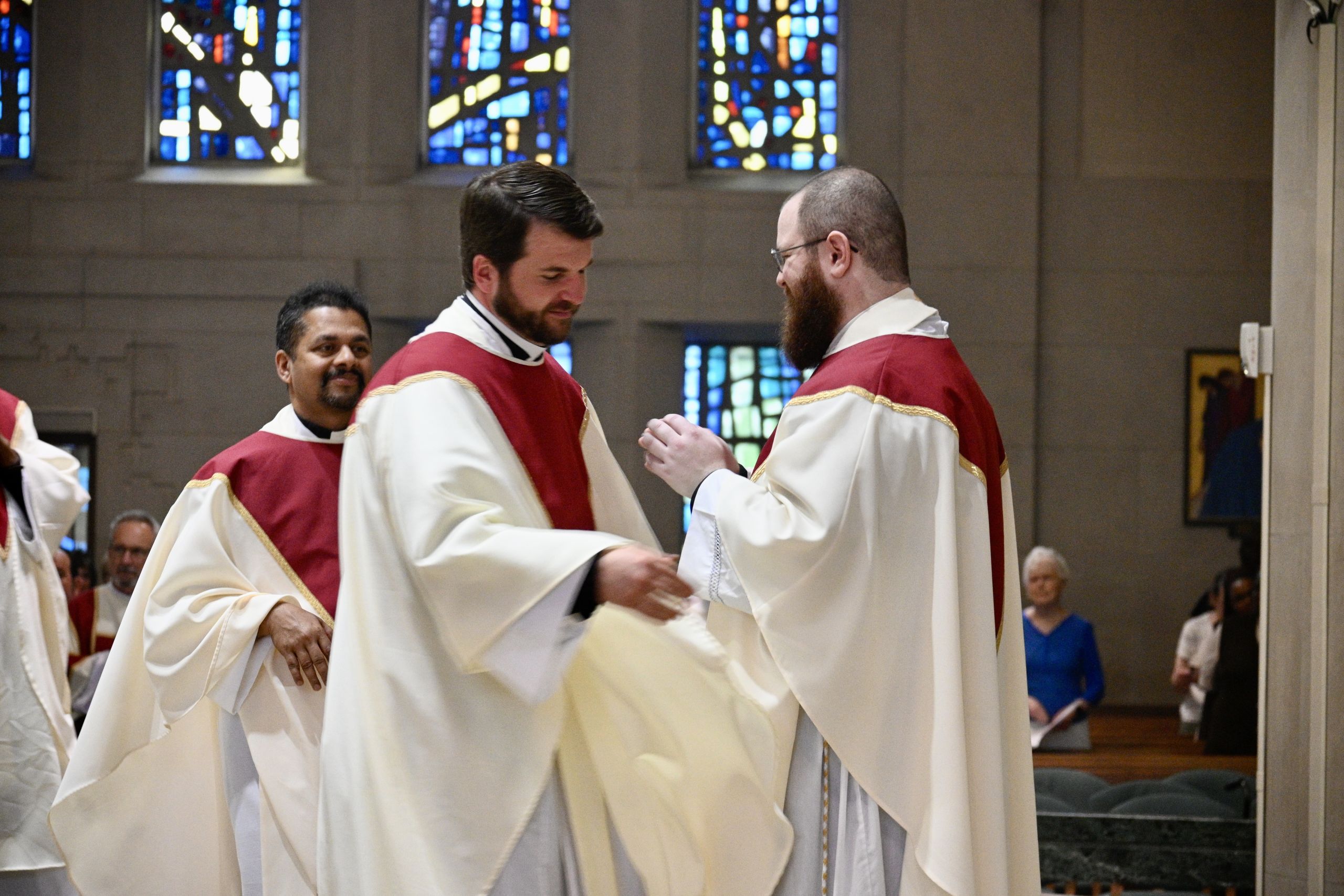 Clergy in white robes with red stoles shake hands during a ceremony inside a church with stained glass windows in the background.