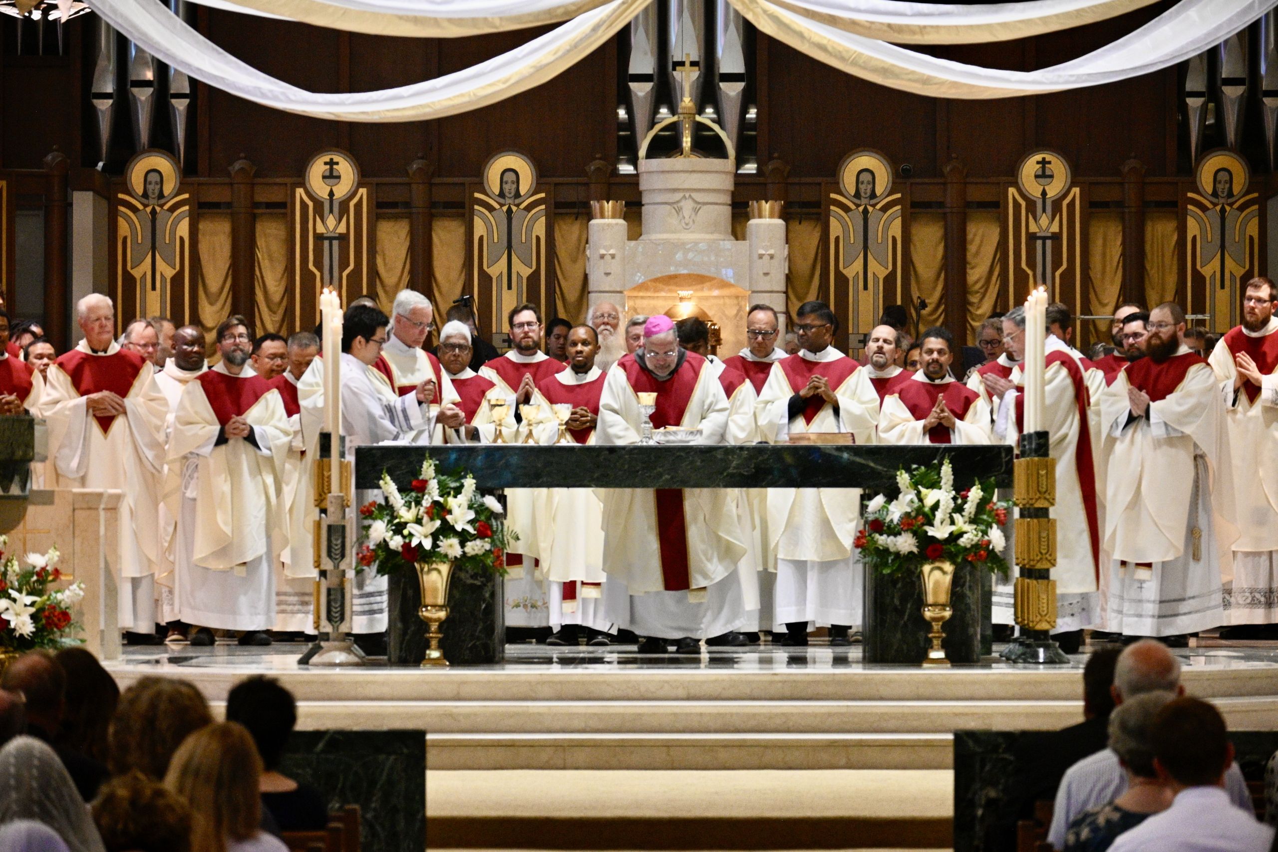 Group of Catholic priests in white and red vestments standing at an altar during a Mass in a richly decorated church downtown-style altar scene.